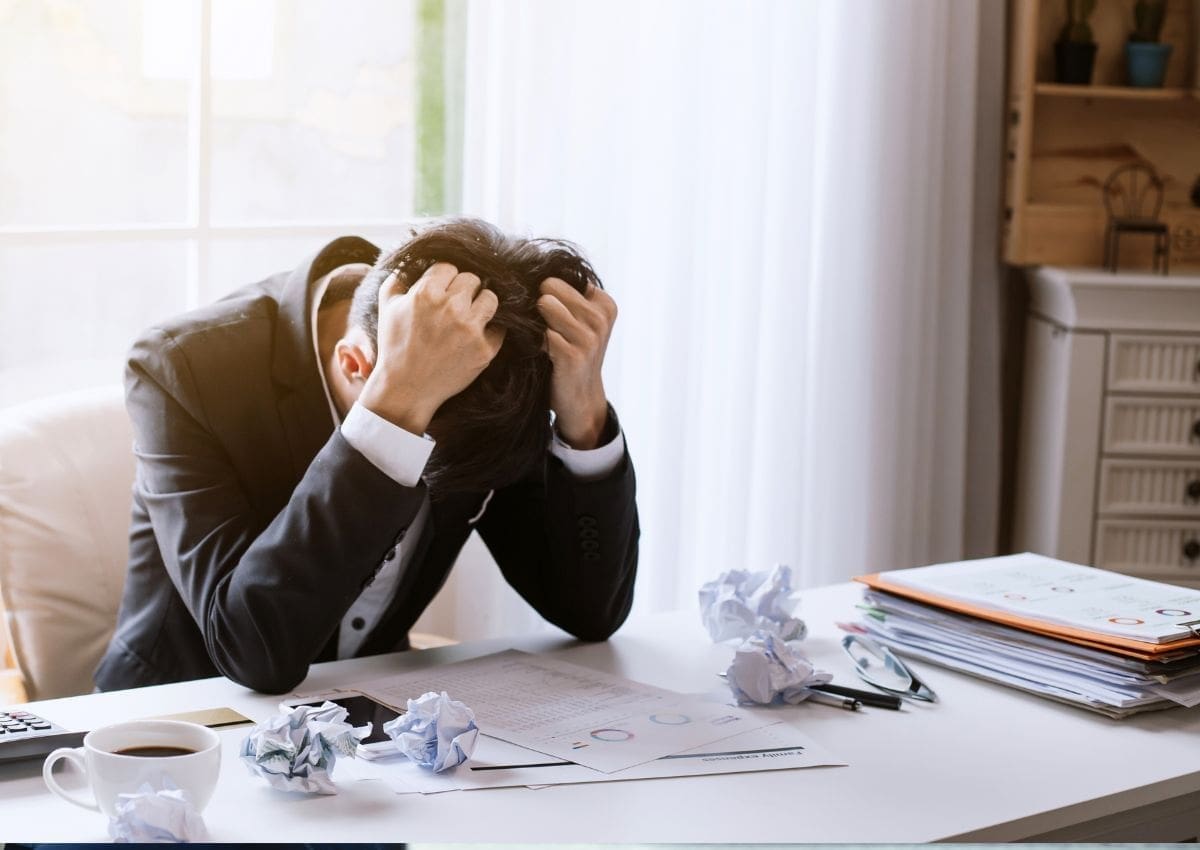 A man in a suit sits at a desk with a frustrated expression, holding his head in his hands amidst crumpled papers and scattered documents.