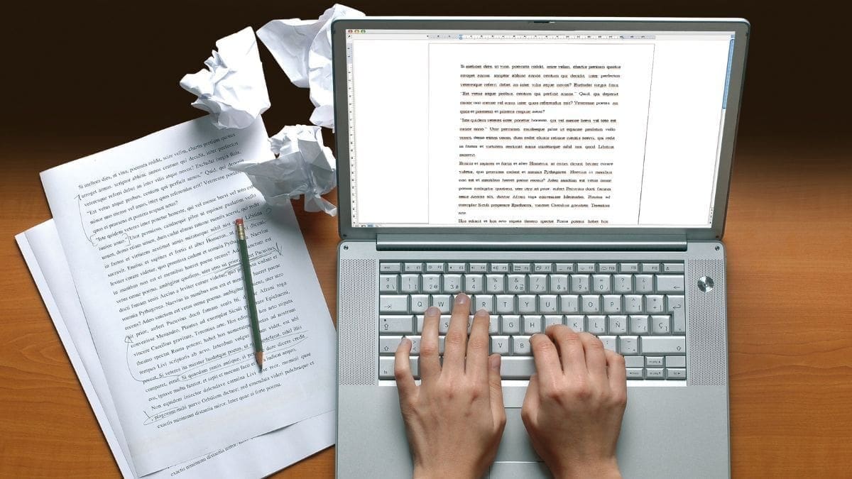 Hands typing on a laptop keyboard with a document on the screen, surrounded by handwritten papers, a pencil, and crumpled drafts.
