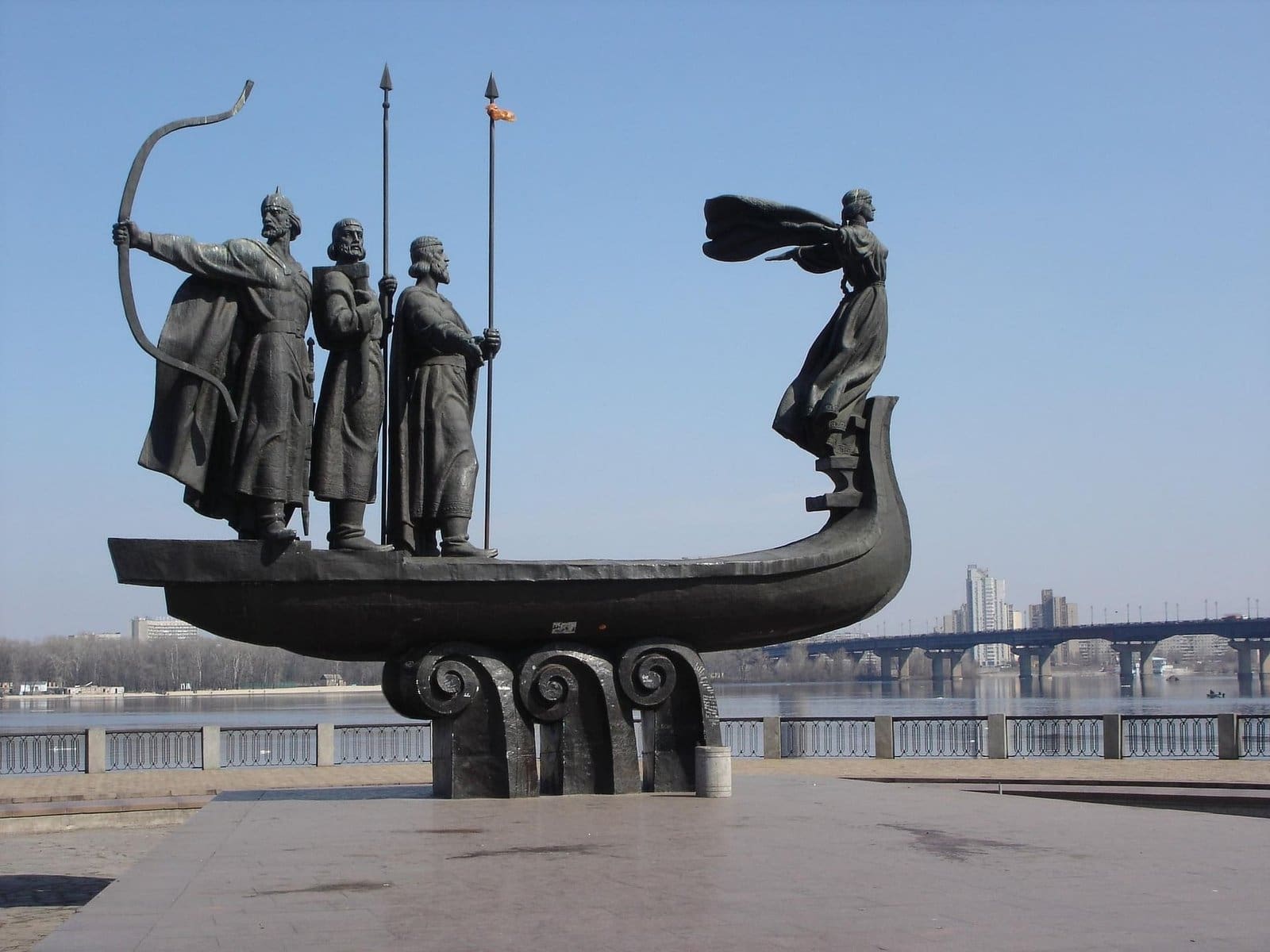 A bronze sculpture of Viking warriors aboard a boat, featuring a female figure in a flowing dress and three male figures, one holding a bow and arrow. The sculpture is set against a clear sky with a city skyline in the background.
