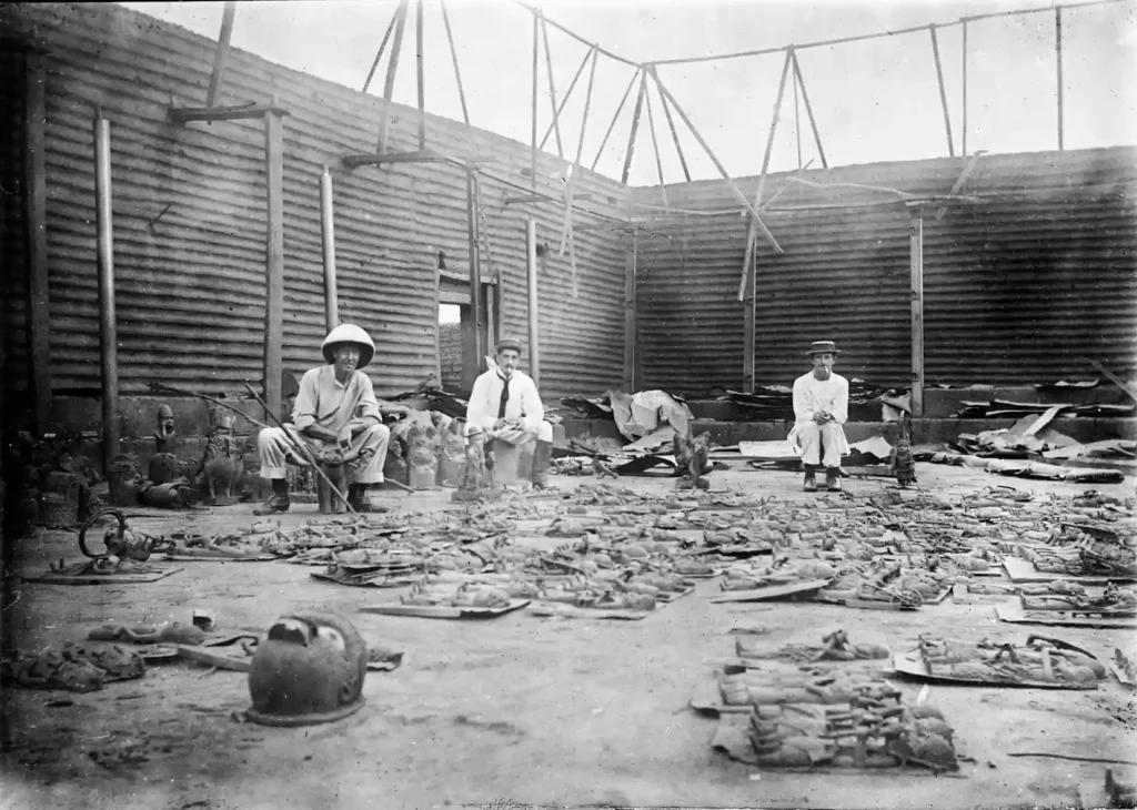 A photograph of the interior of Oba's compound being burnt during the punitive expedition, with bronze plaques in the foreground and three soldiers from the punitive force in the background. Photo: Wikimedia Commons