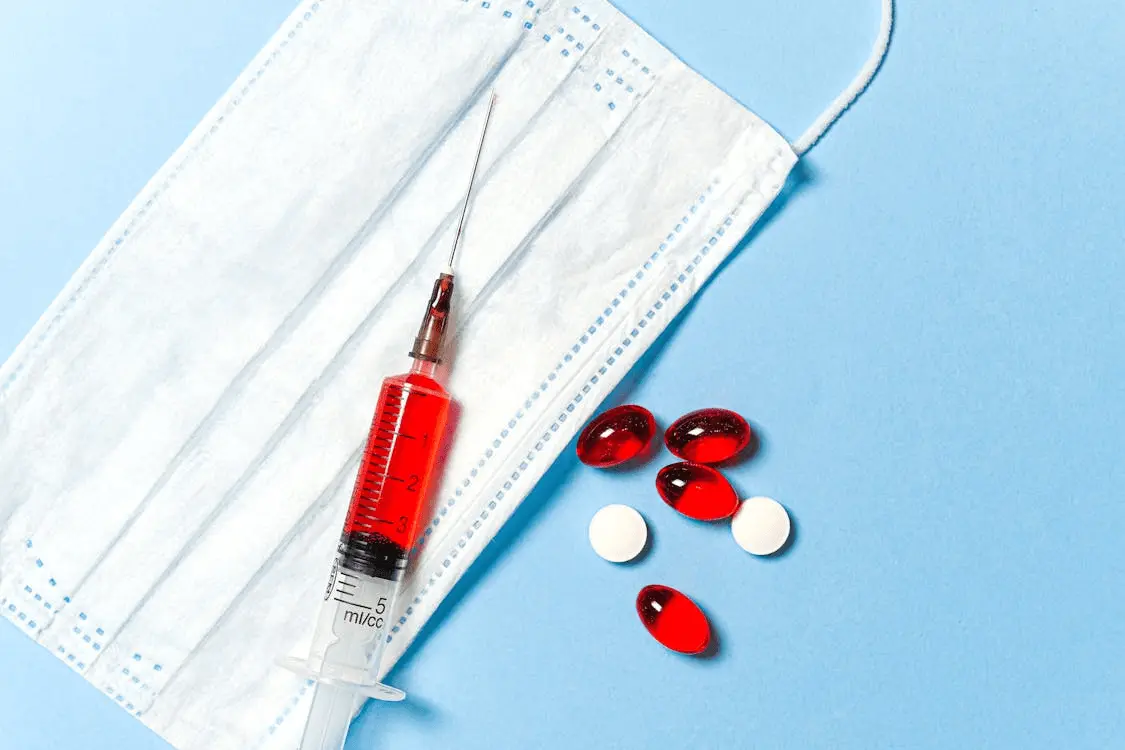 syringe filled with red liquid placed next to a white surgical mask. Also scattered nearby are some red and white capsules. The background is light blue, creating a clean and simple aesthetic