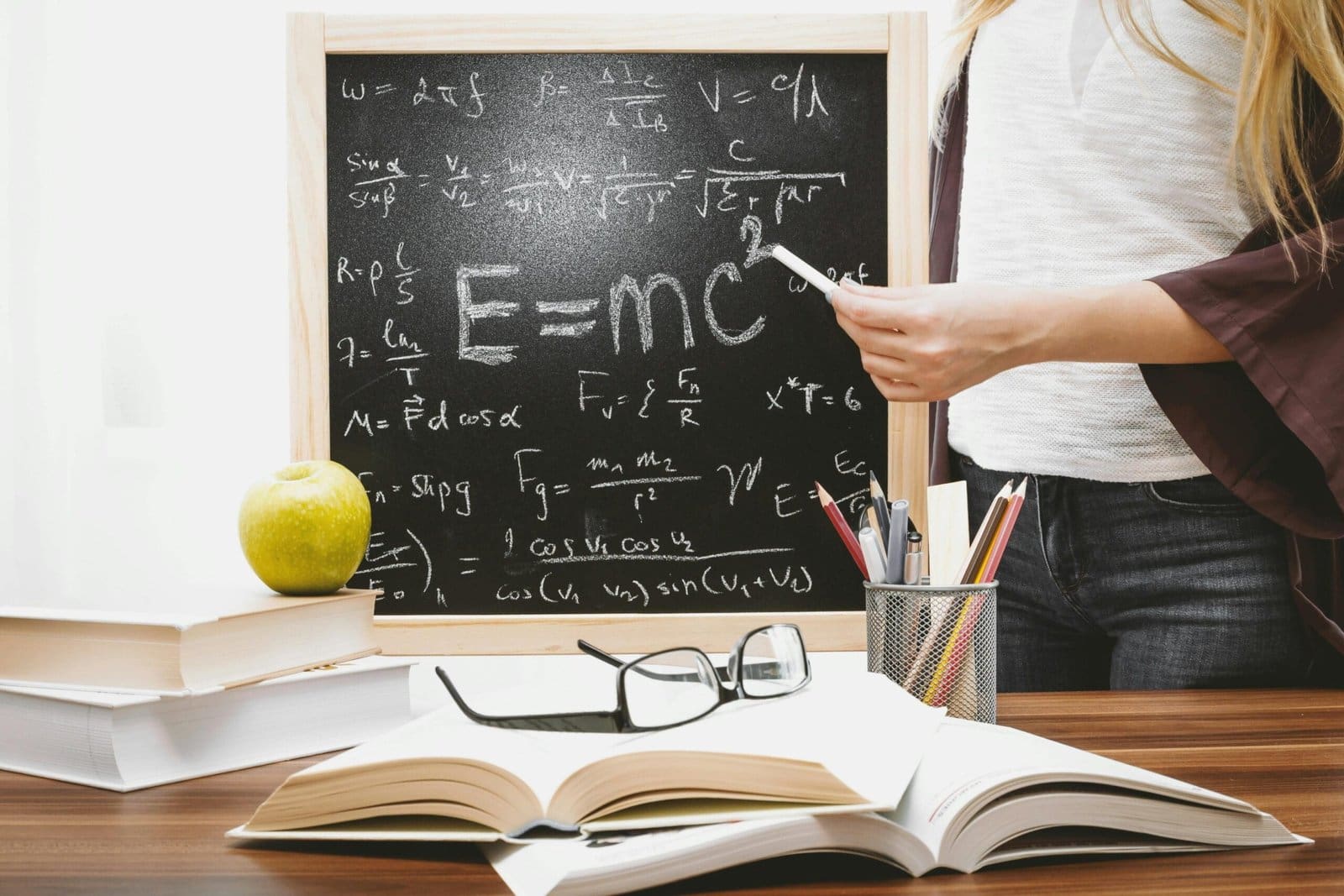 A close-up of a chalkboard with Einstein's famous equation "E=mc²," an apple on the desk, and various school supplies, with a person holding a piece of chalk.