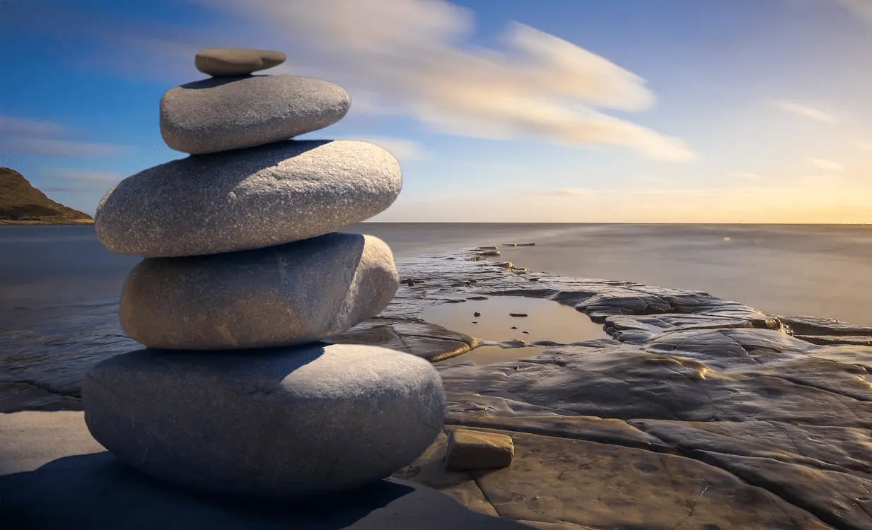 A stack of five smooth, flat stones balanced on a coastal rock platform with the ocean and a golden sunset in the background.