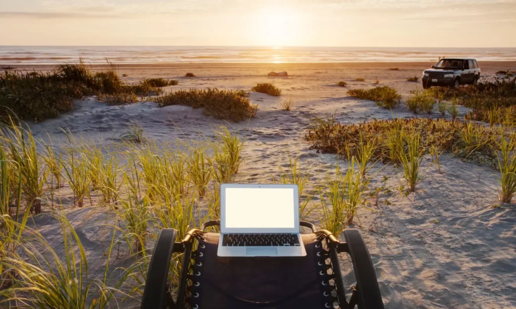 Laptop on a portable chair at a sandy beach with tall grasses, facing a calm horizon at sunset. A rugged vehicle is parked in the background, and the scene conveys a peaceful, travel-ready workspace