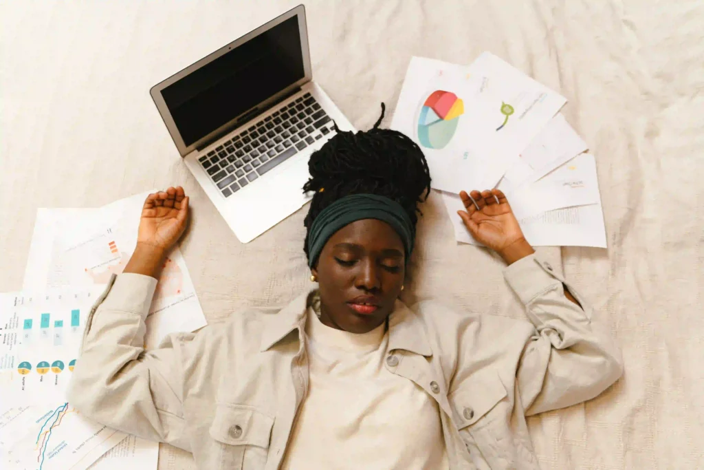 A tired content creator lies on the floor surrounded by her laptop, phone, and notes, eyes closed as she takes a peaceful break from social media and work.