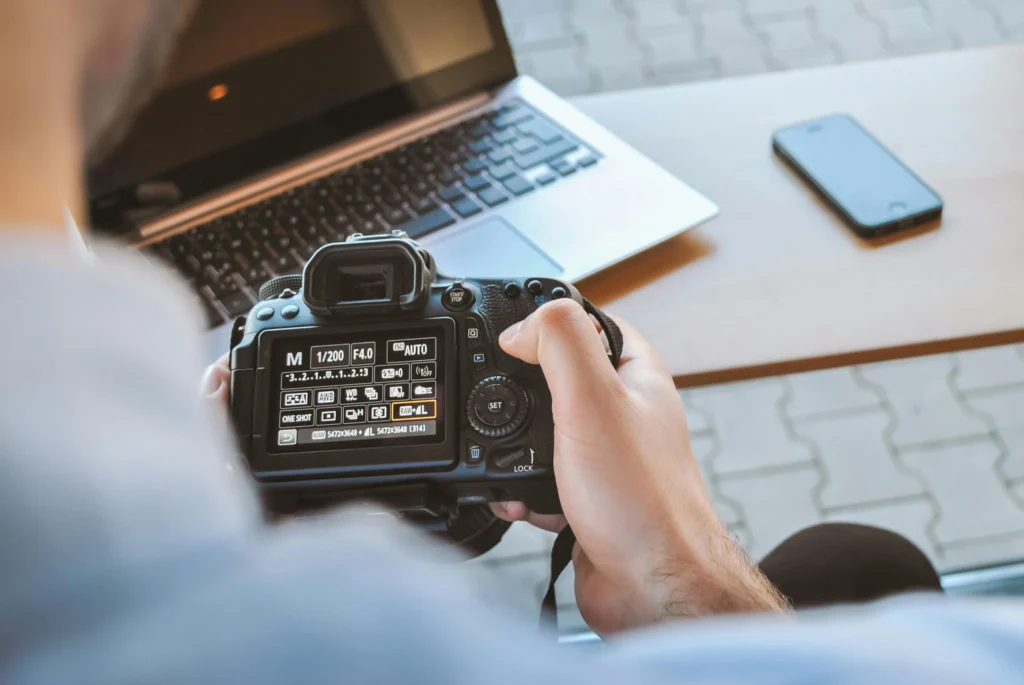 A photographer sits with a DSLR in hand, a laptop and a smartphone on a table nearby, illustrating a travel creator setup focused on power, backup, and file safety.