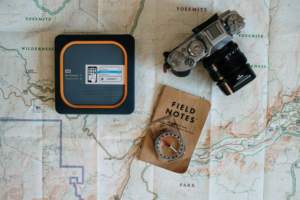 Maps spread out with a camera, a compact tech pack, a field notes notebook, and a compass lying on top.