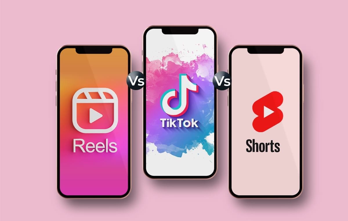 Three smartphones displayed side-by-side on a pink background, comparing the logos and interfaces of the three major short-video platforms: Instagram Reels (left, with its play icon on a gradient background), TikTok (center, with its colorful logo on a white background), and YouTube Shorts (right, with its play button icon on a pale background). 'Vs' is placed between the phones to visually represent the comparison.