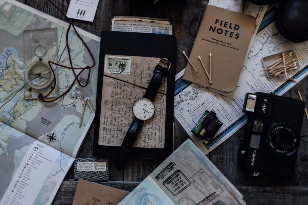 Flat lay of travel essentials including a camera, passport, sunglasses, journal, and smartphone displaying Pinterest boards on a wooden table.